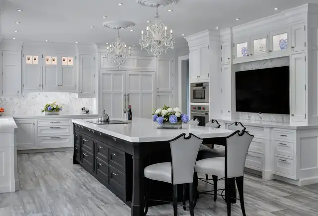 White traditional kitchen with a black island, marble backsplash, crystal chandeliers and a built-in TV wall